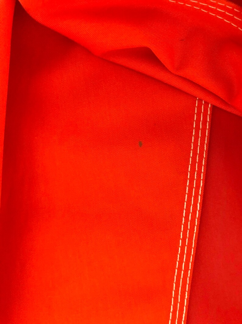 May include: Close-up of a bright orange fabric with white stitching. The fabric has a subtle texture and a small brown stain.