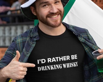 May include: A man wearing a black t-shirt with the text "I'd rather be drinking whisky" printed on it. He is wearing a green and black plaid shirt over the t-shirt and a green and white hat. He is holding a green drink in a clear plastic cup.
