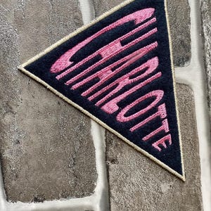 Puede incluir: Parche triangular con el nombre "CHARLOTTE" bordado en rosa sobre fondo azul marino. El parche, con borde crema, está diseñado para aplicar en ropa o accesorios.