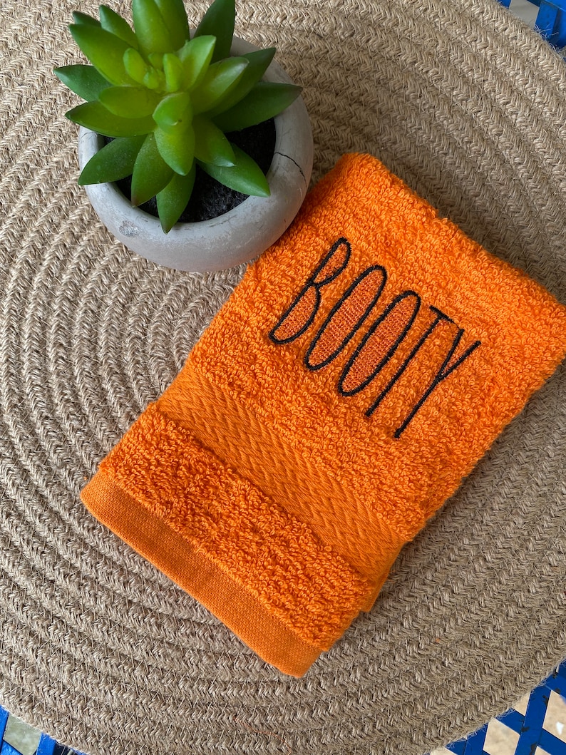 May include: Orange hand towel with the word "BOOTY" embroidered in black thread.