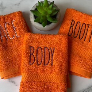May include: Three orange towels with embroidered text. The towels say "FACE", "BODY", and "BOOTY" in white and black thread.