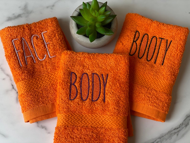 May include: Three orange towels with embroidered text. The towels say "FACE", "BODY", and "BOOTY" in white and black thread.