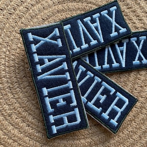 Op de afbeelding: Vier blauwe stoffen patches met witte Romeinse cijfers geborduurd. De patches zijn gestapeld, met de grootste patch onderaan en de kleinste patch bovenaan. De Romeinse cijfers op de patches zijn: "XVII", "XVI", "XV" en "XIV".