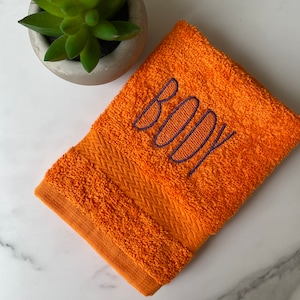 May include: Orange hand towel with the word "BODY" embroidered in purple.
