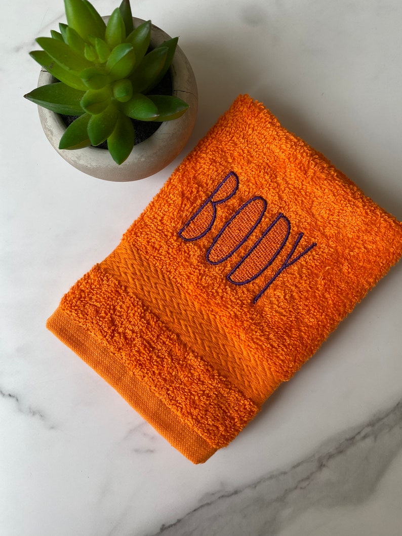 May include: Orange hand towel with the word "BODY" embroidered in purple.