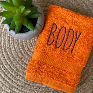 May include: Orange hand towel with the word "BODY" embroidered in purple thread.