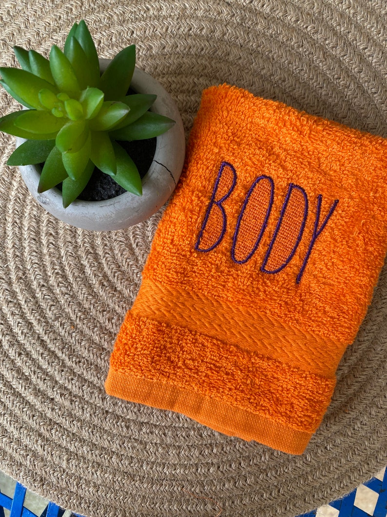 May include: Orange hand towel with the word "BODY" embroidered in purple thread.
