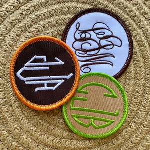 May include: Three embroidered patches with various designs. One is brown with a white stylized letter design and an orange border. Another is white with a brown cursive design and a brown border. The third is tan with a green letter design and a green border.