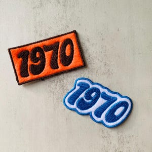 Può includere: Due toppe ricamate con il numero "1970". Una è rettangolare con sfondo arancione e testo marrone. L'altra ha un design blu e bianco con una forma curva.