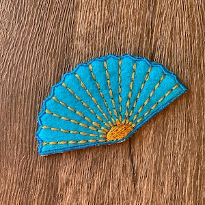 May include: A turquoise fan-shaped felt applique with gold stitching. The fan has a scalloped edge and a sunburst design in the center. The applique is on a brown wooden surface.
