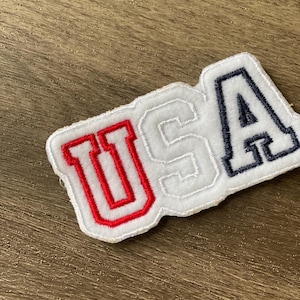 May include: A white, embroidered patch with the word "USA" in red and navy blue lettering. The patch has a white border and is set against a wooden background.