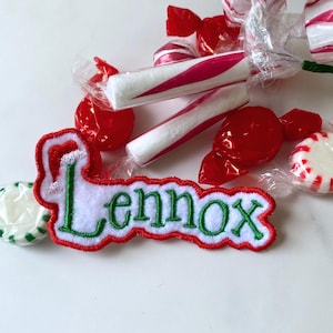 May include: A white felt patch with green and red embroidery that says "Lennox". The patch is surrounded by red and white candy canes and red and white wrapped candies.