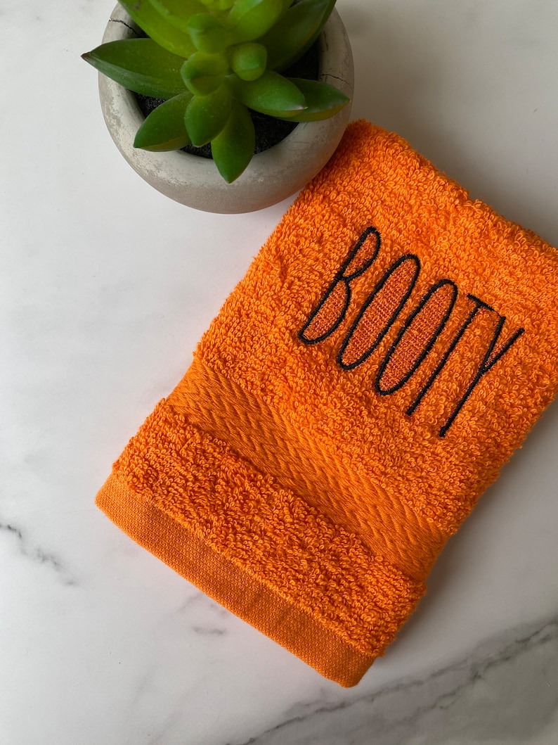 May include: Orange hand towel with black embroidered text that says "BOOTY".