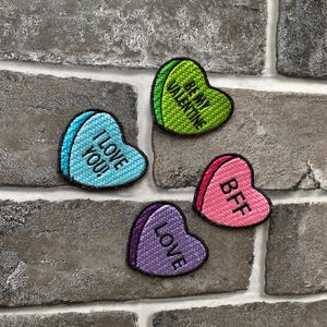 May include: Four heart-shaped patches with embroidered text. The patches are in various colours: light blue with "I LOVE YOU!", green with "BE MY VALENTINE", pink with "BFF", and purple with "LOVE".