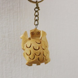 Op de afbeelding: Een houten uil sleutelhanger met een lichtbruine afwerking. De uil heeft grote ogen en een gedetailleerde carving van veren.