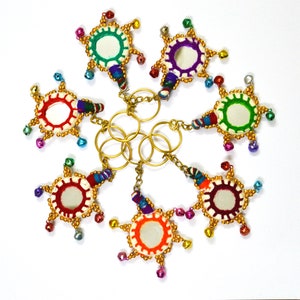 Op de afbeelding: Zeven kleurrijke sleutelhangers met kleine spiegels en gouden accenten. Elke sleutelhanger heeft een andere kleur en een ander patroon van stof en kralen.