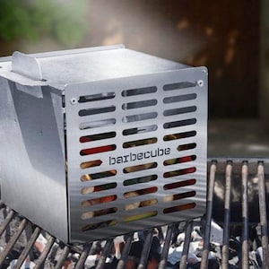 Könnte beinhalten: Ein Edelstahl-Grillwürfel mit dem Wort "barbecube" auf der Seite, der auf einem Grill steht. Der Würfel hat ein geschlitztes Design und enthält sichtbares Gemüse. Der Grill befindet sich über glühenden Kohlen.