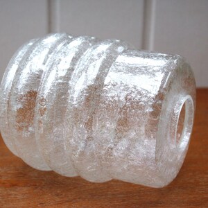 Op de afbeelding: Transparante glazen lampenkap met een getextureerd, geribbeld ontwerp. De cilindrische kap heeft een mat uiterlijk en een cirkelvormige opening aan elk uiteinde. Het glas heeft een bubbelachtige textuur.