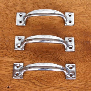 Könnte beinhalten: Drei polierte silberfarbene Metall-Schubladengriffe mit gebogenem Griffdesign. Jeder Griff hat zwei rechteckige Montageplatten mit Schraubenlöchern. Die Griffe sind auf einer Holzoberfläche angeordnet.