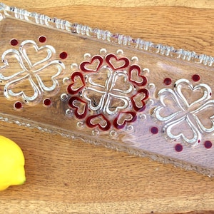Puede incluir: Una bandeja rectangular de vidrio con borde decorativo. La bandeja presenta tres diseños florales hechos de corazones y puntos en vidrio rojo y transparente. Un limón amarillo se encuentra sobre una superficie de madera.