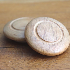Può includere: Due pomelli in legno di colore chiaro con un design circolare. I pomelli sono in legno e hanno una finitura naturale. I pomelli sono su una superficie di legno.