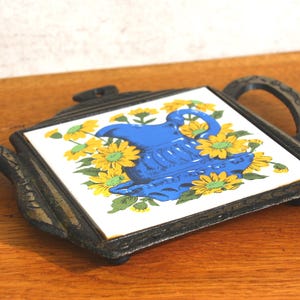 Può includere: Un sottopentola decorativo a forma di teiera. La piastrella centrale presenta una brocca blu circondata da fiori gialli su sfondo bianco. Il sottopentola ha una cornice e un manico in metallo nero.
