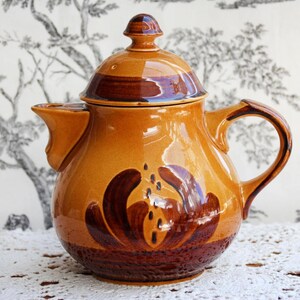 Pode incluir: Uma chaleira de cerâmica com tampa, bico e alça. A chaleira é principalmente laranja com detalhes em marrom escuro e um desenho floral. A chaleira está sobre uma toalha de mesa de renda branca.