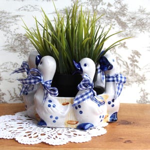 Peut inclure: Un pot en céramique blanc avec des figurines de canards, des accents bleus et des nœuds vichy. Le pot contient de l'herbe verte et repose sur une nappe en dentelle blanche. Le fond présente un motif de toile de Jouy.