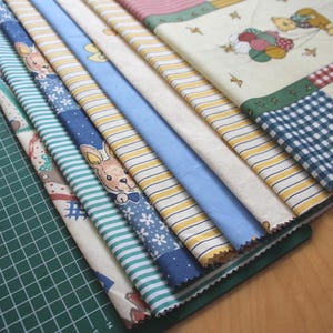 Può includere: Una collezione di campioni di tessuto piegati con vari motivi, tra cui righe gialle e bianche, quadretti vichy blu e bianchi, stampe floreali e un orsetto dei cartoni animati con palloncini su sfondo crema, disposti in una pila diagonale.