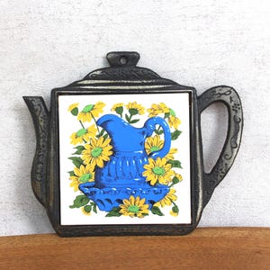 Puede incluir: Un salvamanteles decorativo con forma de tetera. El azulejo central presenta una jarra azul rodeada de flores amarillas. El salvamanteles tiene un marco de metal negro, un asa, un pico y un lazo para colgar.