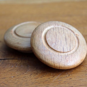 Peut inclure: Deux boutons de meuble en bois clair avec un motif circulaire. Les boutons sont en bois et ont une finition naturelle. Les boutons sont sur une surface en bois.