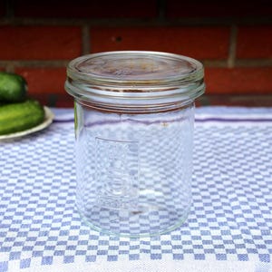 Op de afbeelding: Een heldere glazen pot met deksel, op een blauw-wit geruite tafelkleed. Groene komkommers liggen op een wit bord op de achtergrond. De pot is leeg en klaar voor het inmaken.