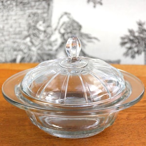 Puede incluir: Una mantequera de cristal transparente con tapa. El plato tiene una base redonda y una tapa abovedada con un asa decorativa. El cristal tiene un diseño acanalado. El plato está sobre una superficie de madera.