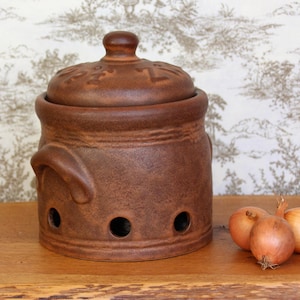 Könnte beinhalten: Ein brauner Keramik-Zwiebeltopf mit Deckel und Belüftungslöchern. Der Topf steht auf einer Holzoberfläche mit drei Zwiebeln daneben.