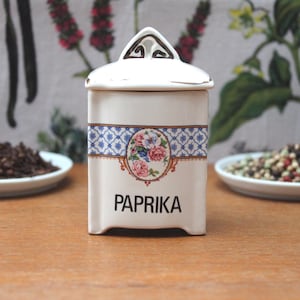 Op de afbeelding: Een witte keramische paprikakruidenpot met een deksel, met een blauw-wit patroonband en een bloemenontwerp. Het woord "PAPRIKA" is in het zwart gedrukt. De pot staat op een houten oppervlak.