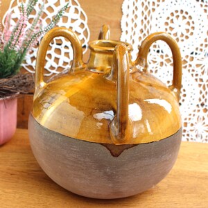 May include: A round ceramic vessel featuring a glossy amber-colored glaze on the upper portion and a matte, textured gray finish on the lower half. The piece includes three vertical handles extending from the shoulder to the neck.
