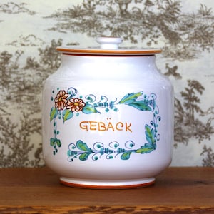 Pode incluir: Jarro de cerâmica branca com tampa, decorado com desenhos florais laranja e verde. O jarro tem a palavra "GEBÄCK" escrita nele.