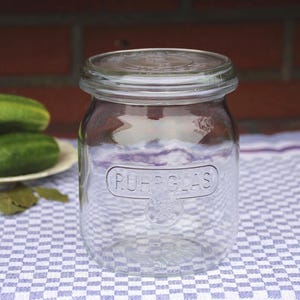 Peut inclure: Un bocal en verre transparent avec un couvercle, étiqueté "RUFF-GLAS", est posé sur un tissu à carreaux bleu et blanc. Des concombres verts sont sur une assiette en arrière-plan. Le bocal est pour la conservation des aliments.