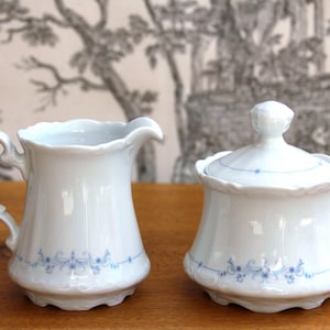 Puede incluir: Un juego de porcelana blanca de jarra para crema y azucarero. La jarra tiene un asa curva y un borde festoneado. El azucarero tiene una tapa con un pomo decorativo. Ambas piezas están adornadas con un delicado patrón floral azul.