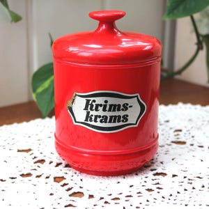 Peut inclure: Un pot rouge vif, cylindrique, avec un couvercle et une petite poignée. Il porte une étiquette blanche et noire avec l'inscription "Krims-krams". Le pot est posé sur une nappe dentelle blanche.