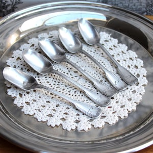 Könnte beinhalten: Sechs silberne Löffel mit verzierten Griffen sind auf einer weißen Serviette auf einem silbernen Tablett angeordnet.