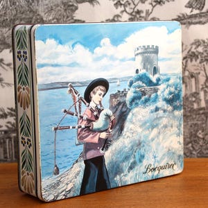 Può includere: Una scatola di latta vintage con un bordo floreale colorato. Il coperchio presenta un dipinto di una persona che suona la cornamusa di fronte a una torre di pietra e un specchio d'acqua. Il testo "Boquirac" è stampato nell'angolo in basso a destra della scatola.