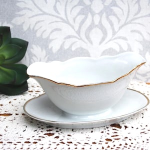 Könnte beinhalten: Weiße Porzellan-Sauciere mit gewelltem Rand und goldenem Rand, auf einer passenden Untertasse. Die Sauciere hat ein klassisches Design, ideal zum Servieren von Saucen. Das Set wird auf einer weißen Spitzendecke präsentiert.