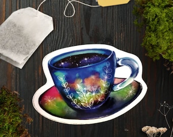 May include: A die-cut sticker featuring a blue teacup and saucer with a galaxy-inspired watercolor design. The cup contains a dark starry sky pattern and is decorated with white line-art botanical sprigs, resting on a colorful saucer.