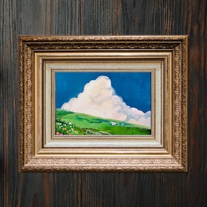 Puede incluir: Pintura de paisaje enmarcada, que representa una colina verde bajo un cielo azul y una gran nube blanca. La obra está en un marco dorado ornamentado, sobre un fondo de madera oscura.