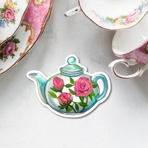 Puede incluir: Una tetera de vidrio transparente con tapa y asa azules, decorada con rosas rosas y hojas verdes.
