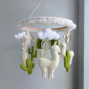 May include: A white and green felt baby mobile with a white cloud, a star, a rainbow, two llamas, and two cacti. The mobile is hanging from a white wooden ring with a white fringe.