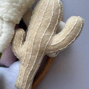 Puede incluir: Un cactus de fieltro beige con detalles cosidos en blanco. El cactus tiene dos brazos y forma parte de una creaci&oacute;n de fieltro m&aacute;s grande. El cactus est&aacute; sobre un fondo gris claro.
