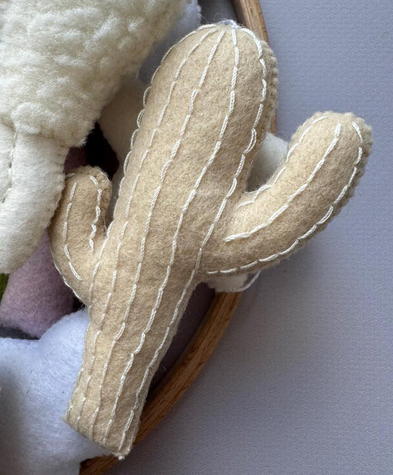 Puede incluir: Un cactus de fieltro beige con detalles cosidos en blanco. El cactus tiene dos brazos y forma parte de una creaci&oacute;n de fieltro m&aacute;s grande. El cactus est&aacute; sobre un fondo gris claro.