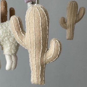 Puede incluir: M&oacute;vil de cactus de fieltro con una paleta de colores neutros. El m&oacute;vil presenta varios cactus de fieltro de diferentes tama&ntilde;os y formas, con detalles de costura blancos. Tambi&eacute;n se ve una llama y otras formas de fieltro.
